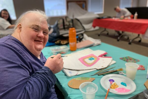 A woman in a purple top looks toward the camera as she holds a paintbrush. On the table in front of her is a tray of paint, brushes and paper on which she has begun painting