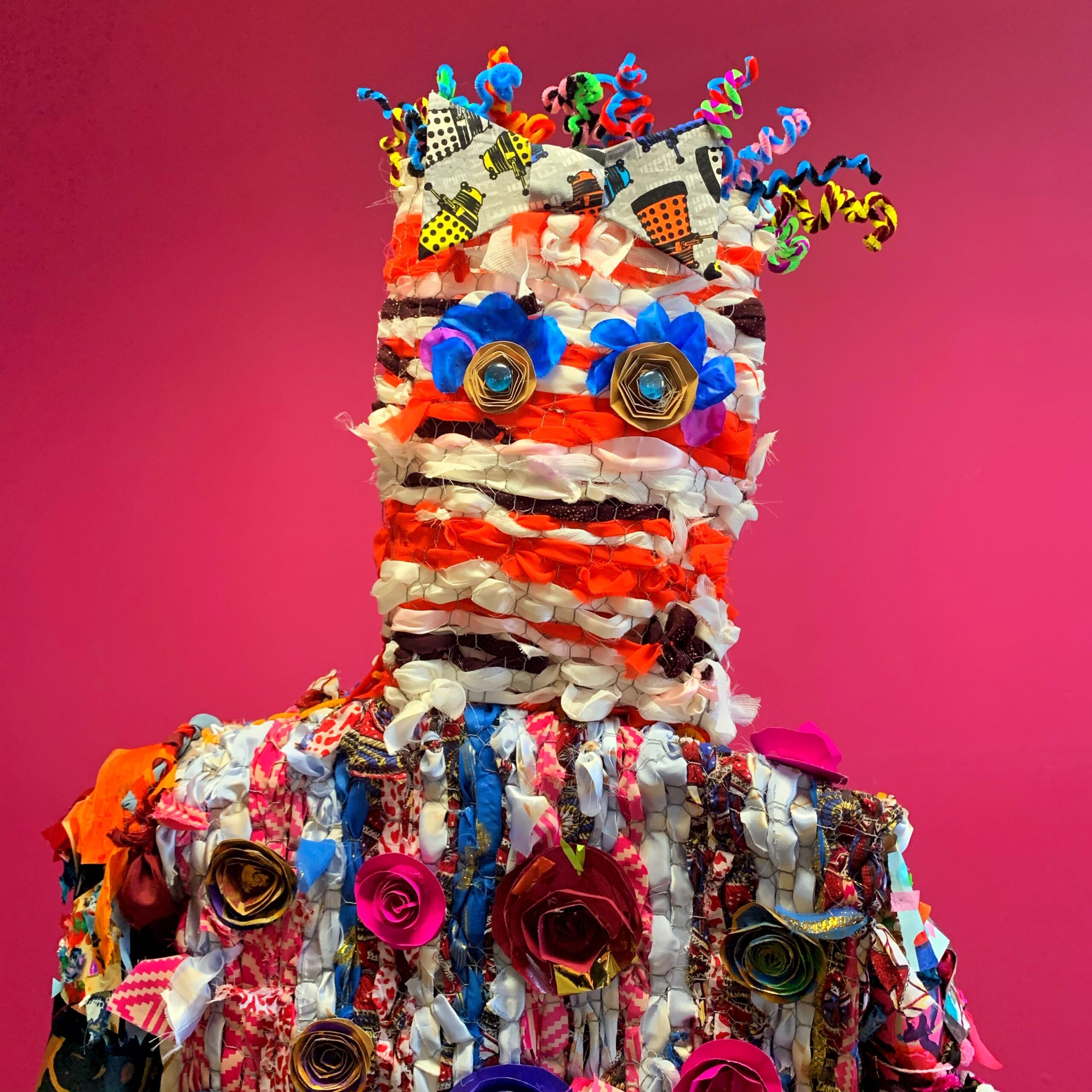This artwork is a figure made of brightly coloured fabric woven through mesh. It is visible from the chest up. It is decorated with paper rosettes , rosettes and marbles for eyes and curly pipe cleaners for hair.